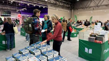 Das Bild zeigt eine Buchmesse oder eine ähnliche Veranstaltung in einer großen, modernen Halle. Der Boden ist mit grauem Teppich ausgelegt. In der Mitte des Bildes stehen mehrere Tischgruppen mit grünen Tischdecken, auf denen viele Bücher displayed sind.   Vorne im Bild stehen zwei Personen im Gespräch. Die linke Person hat einen Rucksack auf dem Rücken und trägt ein kariertes Hemd, während die rechte Person eine hellblaue Jacke und einen Schal trägt. Neben ihnen ist eine dritte Person, die eine rote Bluse trägt und auf einen Tisch zeigt, auf dem Bücher liegen.  Im Hintergrund sind weitere Besucher der Veranstaltung zu sehen, einige durchstöbern die Buchauslagen, während andere in Gespräche vertieft sind. An der Wand hängen große Banner oder Werbetafeln, und es gibt einige Ballons. Das Licht in der Halle ist hell und es gibt technische Installationen an der Decke.