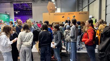 Das Bild zeigt eine große Menschenmenge in einer Messehalle. Im Vordergrund sind mehrere Kinder und Jugendliche zu sehen, die in einer Reihe stehen und sich unterhalten. Einige von ihnen tragen bequeme Kleidung, wie Sweatshirts und weite Hosen.   Hinter ihnen steht ein größerer Stand mit hellbraunen Wänden, der mit der Aufschrift „RÜCKZUGSORT MITTEN IM ALLTAG“ versehen ist.   Im Hintergrund sind viele weitere Personen sichtbar, die durch die Halle gehen oder sich um verschiedene Stände gruppieren. Die Beleuchtung ist teilweise bunt, was auf eine lebhafte Atmosphäre hinweist.   Die Halle hat hohe Wände und trägt den typischen Charakter von Messeveranstaltungen. Es sind keine spezifischen Orte oder Gebäude außerhalb der Messe zu erkennen.