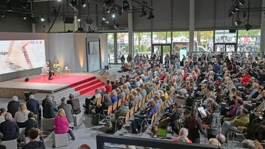 Auf einer Bühne sitzen 2 Personen und diskutieren vor großem Publikum. 