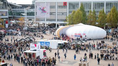 Vor der Messehalle der Frankfurter Buchmesse sind Container und ein großes ovales Zelt aufgebaut, um die sich Menschenmassen bewegen.