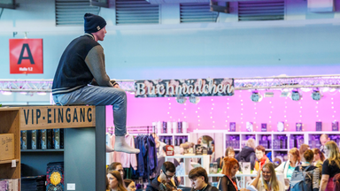 Ein Mann sitzt oben auf einer Stellwand zwischen Buchmessen-Gängen und Ständen über dem Menschengewusel.