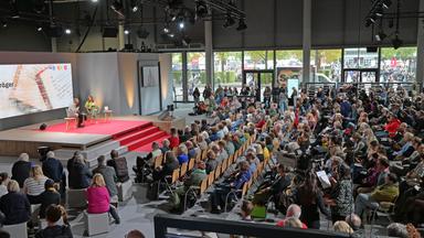 In einem großen Saal sitzen Menschen in Stuhlreihen vor einer Bühne, auf der auf einem rote  Teppich zwei Personen sitzen und sich unterhalten.