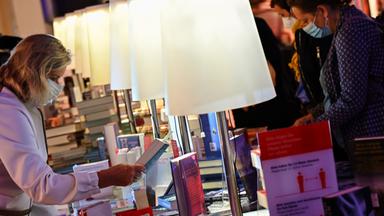 Menschen mit medizinischen Gesichtsmasken stehen um einen langen Tisch mit vielen Büchern und Lampen und schauen die Bücher an.