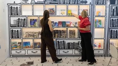 Zwei Frauen räumen ein Bücherregal ein, das wie ein gemalter Bücherschrank aussieht.