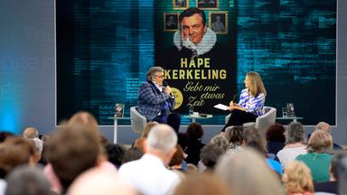 Ein Mann und eine Frau sitzen auf einer Bühne vor Publikum und unterhalten sich. Im Hintergrund sieht man ein großformatiges Buch.
