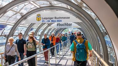 Menschen laufen durch einen mit Glas überdachten Gang auf der Leipziger Buchmesse. Über ihnen weist ein Banner auf die Buchmesse 2024 hin.