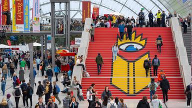 Menschen laufen auf einer großen Treppe, deren Stufen mit dem Logo der Leipziger Buchmesse, einem großen gelben Buch unter einem riesigen Auge mit Krone, beklebt sind.