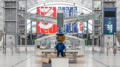 Leere Halle auf der Buchmesse Leipzig mit Messe-Maskottchen und Bannern von 3sat und ARD-Sendern.