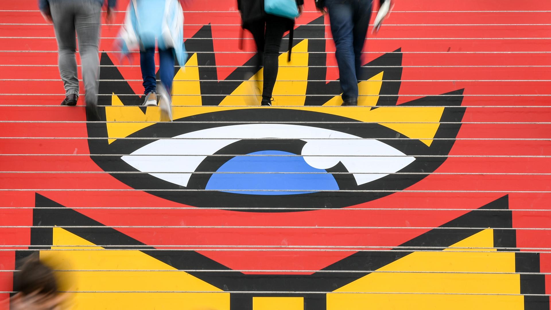 Menschen laufen auf der Buchmesse Leipzig über eine Treppe, auf der das Logo der Buchmesse, ein großes buntes stilisiertes Auge zu sehen ist.