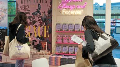 Zwei junge Frauen mit Taschen über der Schulter beugen sich über ausgelegte Bücherstapel mit bonbonfarbenen Schnitten. An der Wand ist in pinken Neon-Röhren-Buchstaben zu lesen: Love wins always and forever.