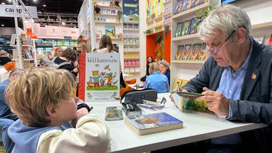 Ein Mann sitzt an einem Büchertisch und hält ein Buch in den Händen, ihm gegenüber leht sich ein kleiner Junge auf den Tisch und sieht ihm zu. Beide sind umgeben von Stellwänden, auf denen Bücher präsentiert werden.