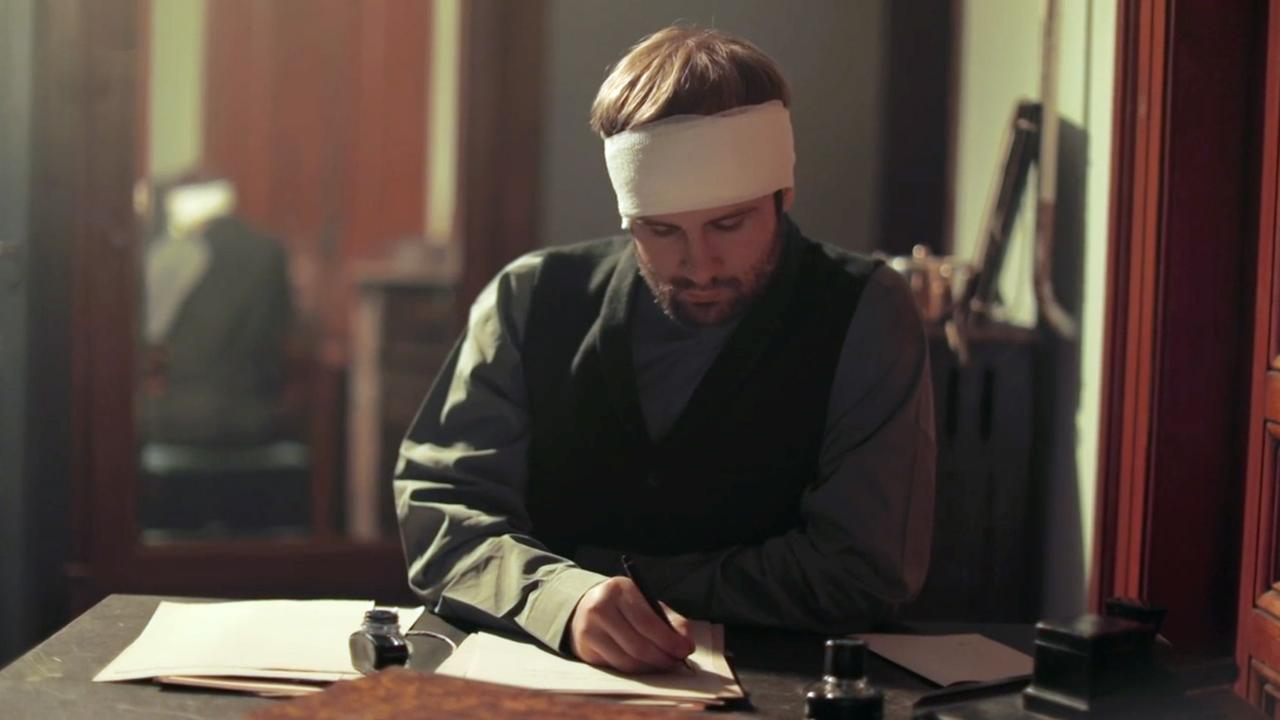 Szene aus "Samson und Nadjeschda" von Andrej Kurkow. Ein Mann mit einem Kopfverband sitzt an einem Tisch und schreibt in einem dämmrigen Raum. Mehrere Papiere und Tintenfässer stehen auf dem Tisch.