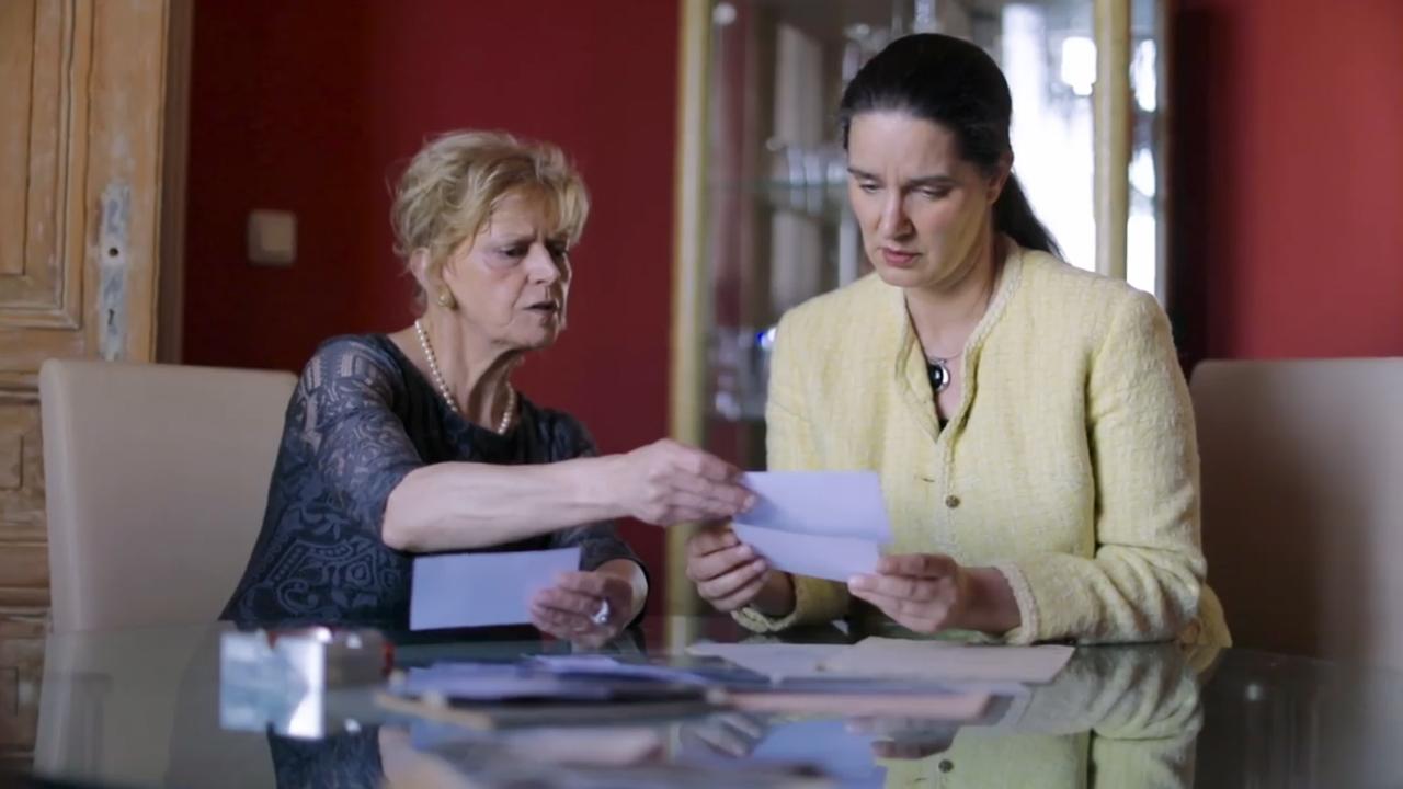 Szene aus "Die Postkarte" von Anne Berest: Eine ältere und jüngere Frau sitzen an einem Tisch und schauen sich verschiedene Dokumente an.