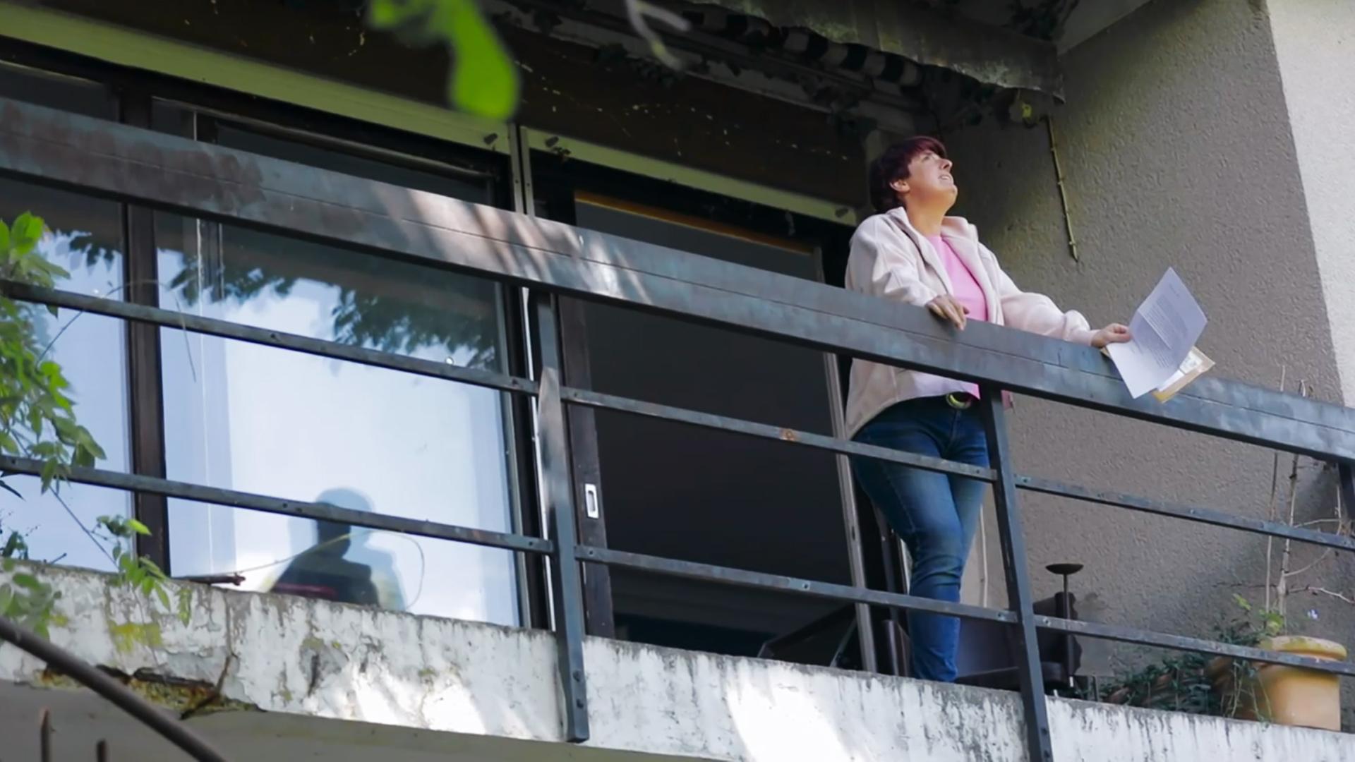 Szene aus "Bevor ich es vergesse" von Anne Pauly. Eine Frau steht auf einem Balkon und hält einen Brief in der Hand. Sie blickt in den Himmel und wirkt traurig.