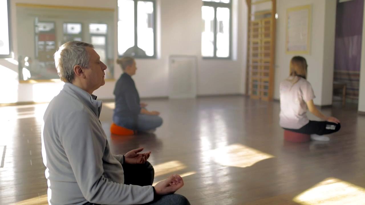 Szene aus "Yoga" von Emmanuel Carrère. Mehrere Personen sitzen in einem großen Raum im Schneidersitz auf einem Yogakissen und meditieren.