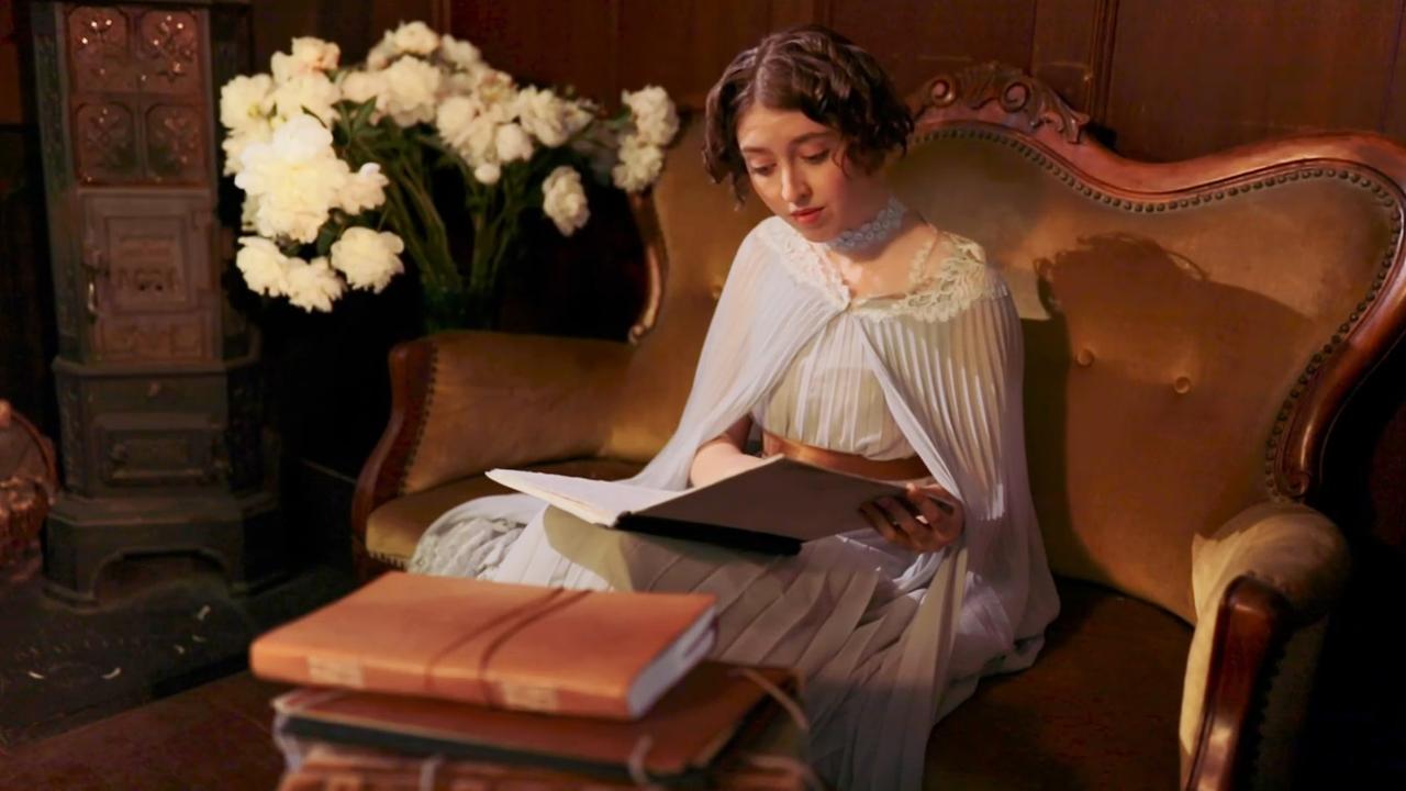 Szene aus "Reise ohne Wiederkehr" von David Diop. Eine Frau mit weißem Spitzenkleid sitzt auf einem grünen Sofa. Auf ihrem Schoß liegt ein aufgeschlagenes Buch indem sie vertieft liest. Links neben ihr steht ein weißes Blumenarrangement auf einem Beistelltisch. Vor dem Sofa steht ein Couchtisch, auf dem ledergebundene Aufzeichnungsmappen liegen.