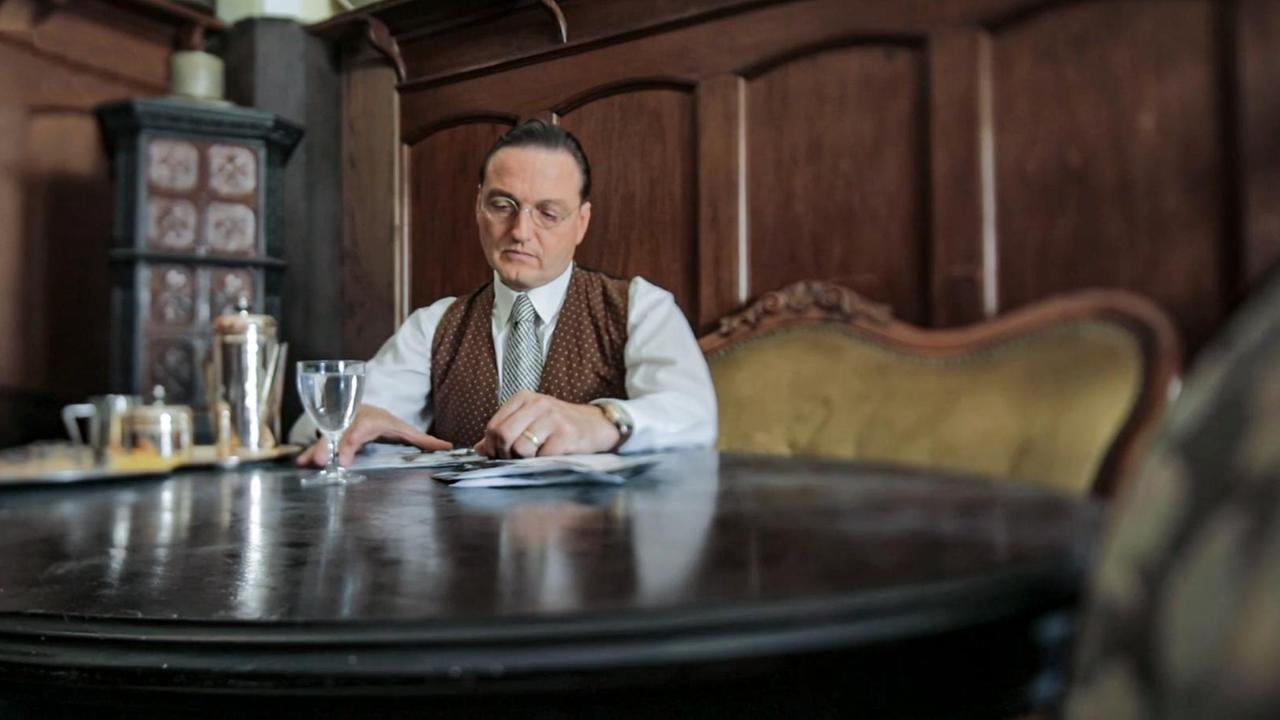 Szene aus "Lichtspiel" von Daniel Kehlmann. Ein Mann mit Brille sitzt an einem runden, dunklen Tisch. Vor ihm liegen Fotografien auf dem Tisch, die er nachdenklich betrachtet. Auf dem Tisch steht eine Kaffeekanne und -tasse auf einem Tablett und ein Glas Wasser.