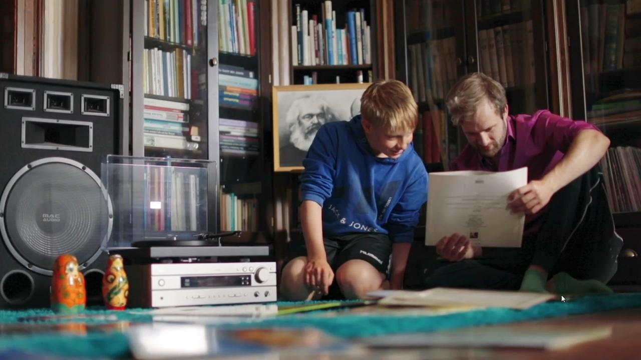 Szene aus der filmischen Buchempfehlung "Die Lüge" von Mikita Franko. Ein Mann und ein Kind sitzen auf dem Fußboden. Der Mann hält ein Schallplattencover in den Händen, welches er sich mit dem Jungen betrachtet. Links von den beiden steht eine Anlage und Box auf dem Boden. Im Hintergrund befinden sich Bücherregale. 