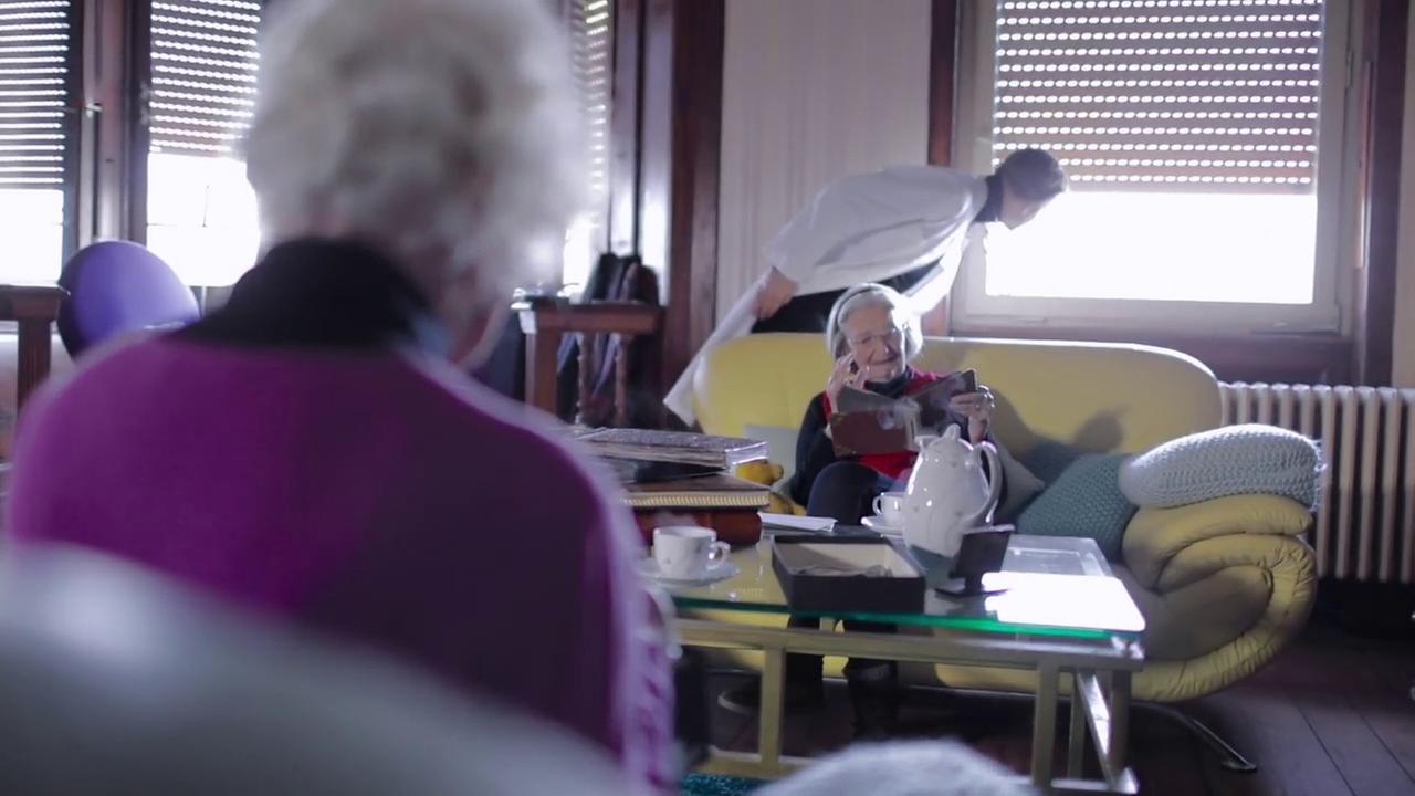 Szene aus "Zeitzuflucht" von Georgi Gospodinov. Zwei ältere Frauen sitzen jeweils auf einem Sofa. In der Mitte steht ein Couchtisch aus Glas, auf dem ein weißes Teeservice mit zwei Teetassen und eine offene schwarze Schachtel stehen. Eine Frau sitzt mit dem Rücken zum Betrachter und trägt einen lila Pulli. Im Hintergrund steht ein gebückter Mann im weißen Artzkittel und blickt aus dem Fenster.