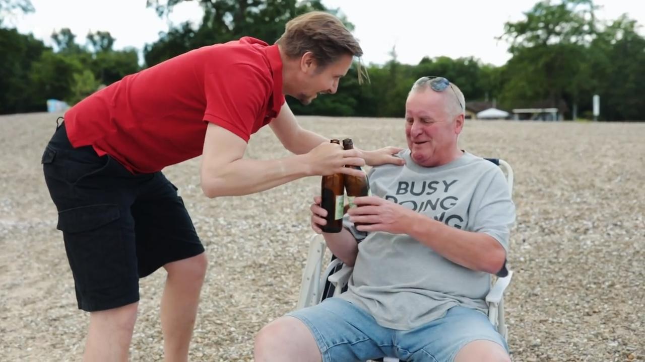 Szene aus "Ein Sommer in Niendorf" von Heinz Strunk. Ein Mann mit grauen T-Shirt und kurzer Jeans sitzt in einem Sonnenstuhl am Strand. Neben ihm beugt sich ein Mann mit rotem Poloshirt und kurzer schwarzer Hose zu ihm. Seine linke Hand liegt auf der Schulter des sitzenden Mannes. Seine rechte Hand umfasst zwei Bierflaschen, die er dem sitzenden Mann anreicht. 