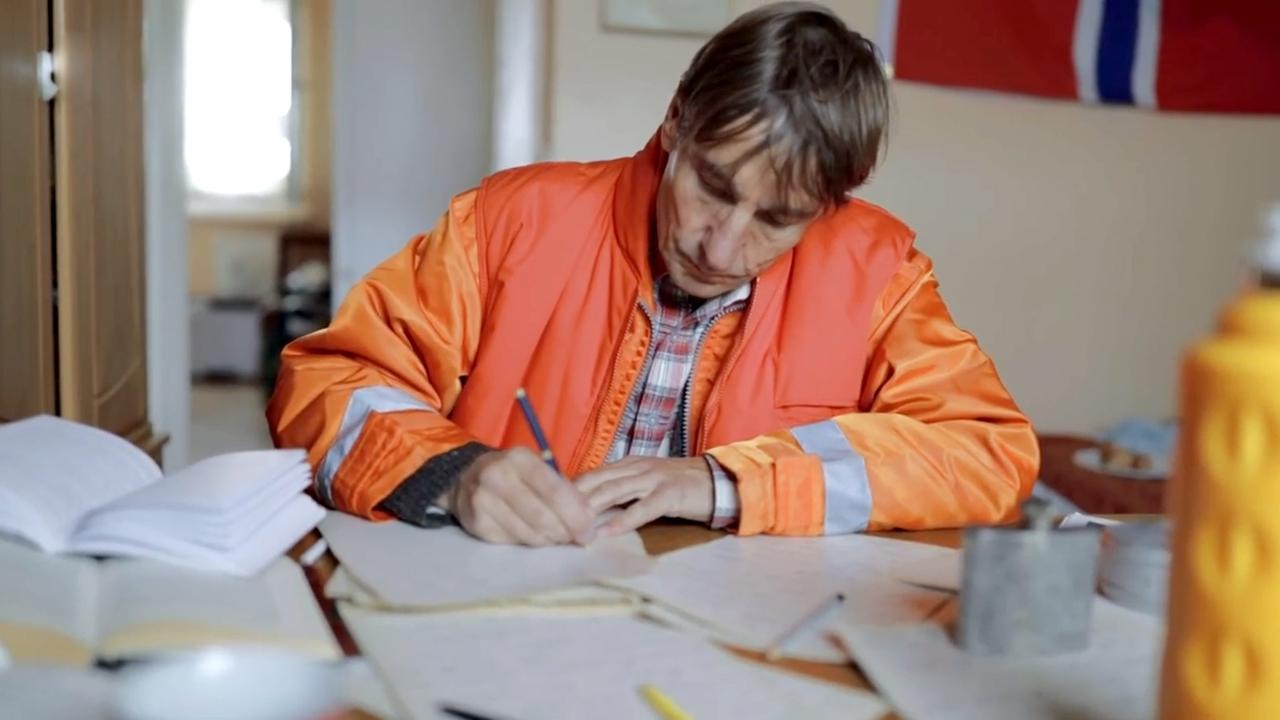 Szene aus "Auf frischer Tat" von Johan Harstad. An einem Schreibtisch sitzt ein Mann mit einer organgefarbenen Jacke und schreibt. Zahlreiche beschriebene Blätter und Bücher sind auf dem Tisch verteilt.