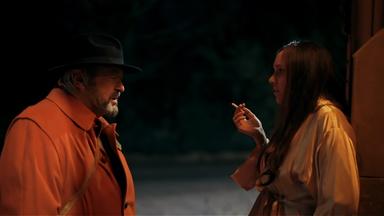 Szene aus "Singularitäten" von John Banville. Ein älterer Mann und eine junge, rauchende Frau stehen sich im Dunkeln im Freien gegenüber und sehen sich an. Der Mann steht links und trägt einen rostfarbenen Mantel und einen schwarzen Hut. Er hat einen grau melierten Vollbart. Die junge Frau steht rechts, trägt einen beigefarbenen Mantel und hält in der rechten Hand eine brennende Zigarette. Sie hat braune, lange Haare.