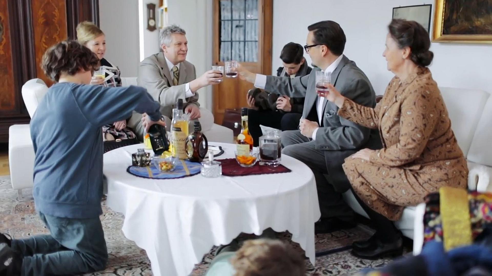 Szene aus "Die Netanjahus" von Joshua Cohen: Zwei Ehepaare sitzen auf zwei Sofas, die um einen Couchtisch mit weißer Decke gruppiert sind. Sie stoßen mit gefüllten Gläsern an. Auf dem Tisch stehen verschiedene Getränkeflaschen. Im Vorder- und Hintergrund sind Kinder zu sehen.