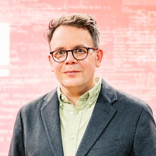 Eine Person mit Brille steht vor einem rosa-roten Hintergrund mit dem weißen Schriftzug „BUCHZEIT“. Sie trägt ein dunkelblaues Oberteil. Das Porträt entstand im Rahmen der Frankfurter Buchmesse 2025 während der Aufzeichnung der Literatursendung „Buchzeit“ auf der Bühne von ARD, ZDF und 3sat.