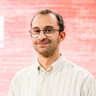 Ein Mann mit Brille steht vor einem rosa-roten Hintergrund mit dem weißen Schriftzug „BUCHZEIT“. Er trägt ein gestreiftes Hemd. Das Porträt wurde während der Frankfurter Buchmesse 2025 aufgenommen, im Rahmen der Aufzeichnung der Literatursendung „Buchzeit“ auf der Bühne von ARD, ZDF und 3sat.