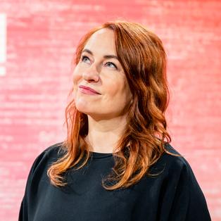 Eine Person mit langen, gewellten Haaren steht vor einem rot-pink strukturierten Hintergrund mit dem weißen Schriftzug „BUCHZEIT“. Sie trägt ein dunkles Oberteil und blickt leicht nach oben. Das Porträt entstand im Rahmen der Frankfurter Buchmesse 2025 während der Aufzeichnung der Literatursendung „Buchzeit“ auf der Bühne von ARD, ZDF und 3sat. 