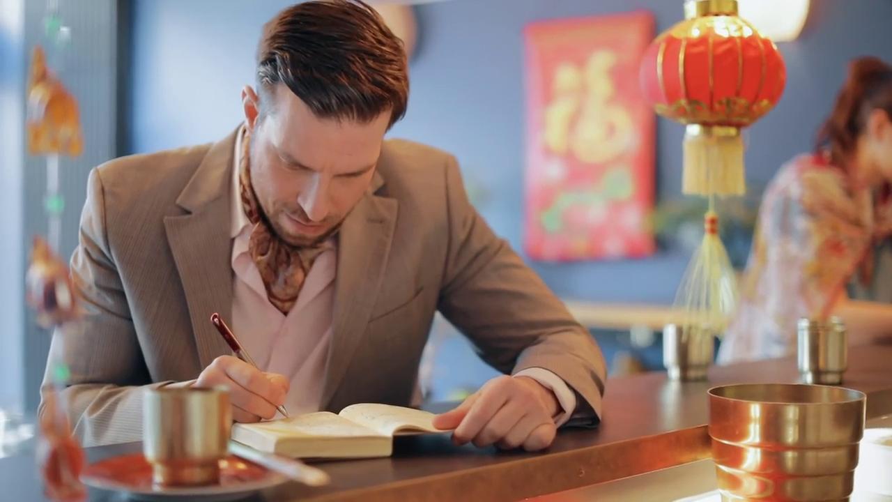 Szene aus "Mr. Woolf" von Jan Kjærstad. Ein jüngerer Mann in einem feinen beigen Anzug sitzt an einer Bar in einem chinesischen Restaurant. Er schreibt in ein Notizbuch.