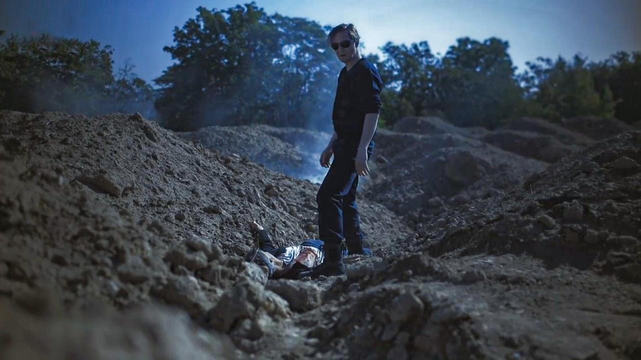 Szene aus "Hund 51" von Laurent Gaudé. Eine Leiche liegt auf dem erdigen Boden. Ein schwarz gekleideter Mann mit Sonnenbrille steht rechts neben der Leiche.