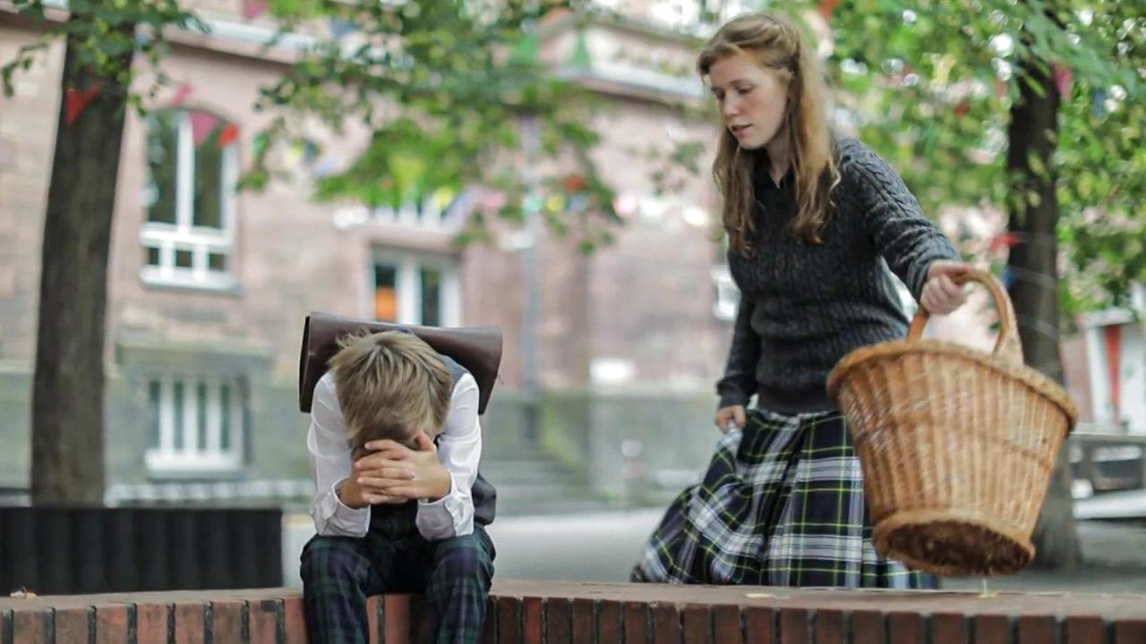 Szene aus "Übertretung" von Louise Kennedy. Ein Junge mit Schulranzen auf dem Rücken sitzt auf einer Mauer. Er hat seinen Kopf gesenkt, seine Arme sind auf den Beinen abstützt und seine Hände vor Gesicht verschränkt. Rechts von ihm steht eine junge Frau mit einem Weidenkorb.