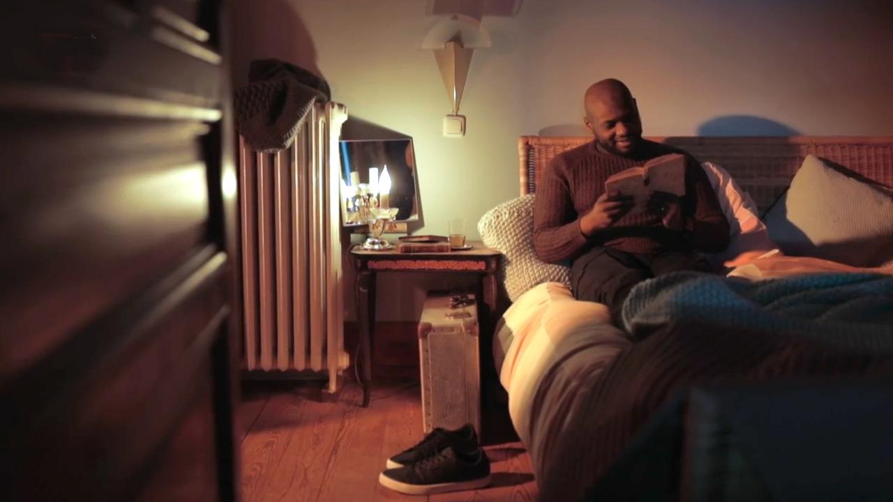 Szene aus "Die geheimste Erinnerung der Menschen" von Mohamed Mbougar Sarr: Ein Mann sitzt im Bett und liest interessiert in einem Buch. Neben dem Bett steht ein Nachtisch mit einer brennenden Lampe. Seine Turnschuhe stehen seitlich vor dem Bett. 