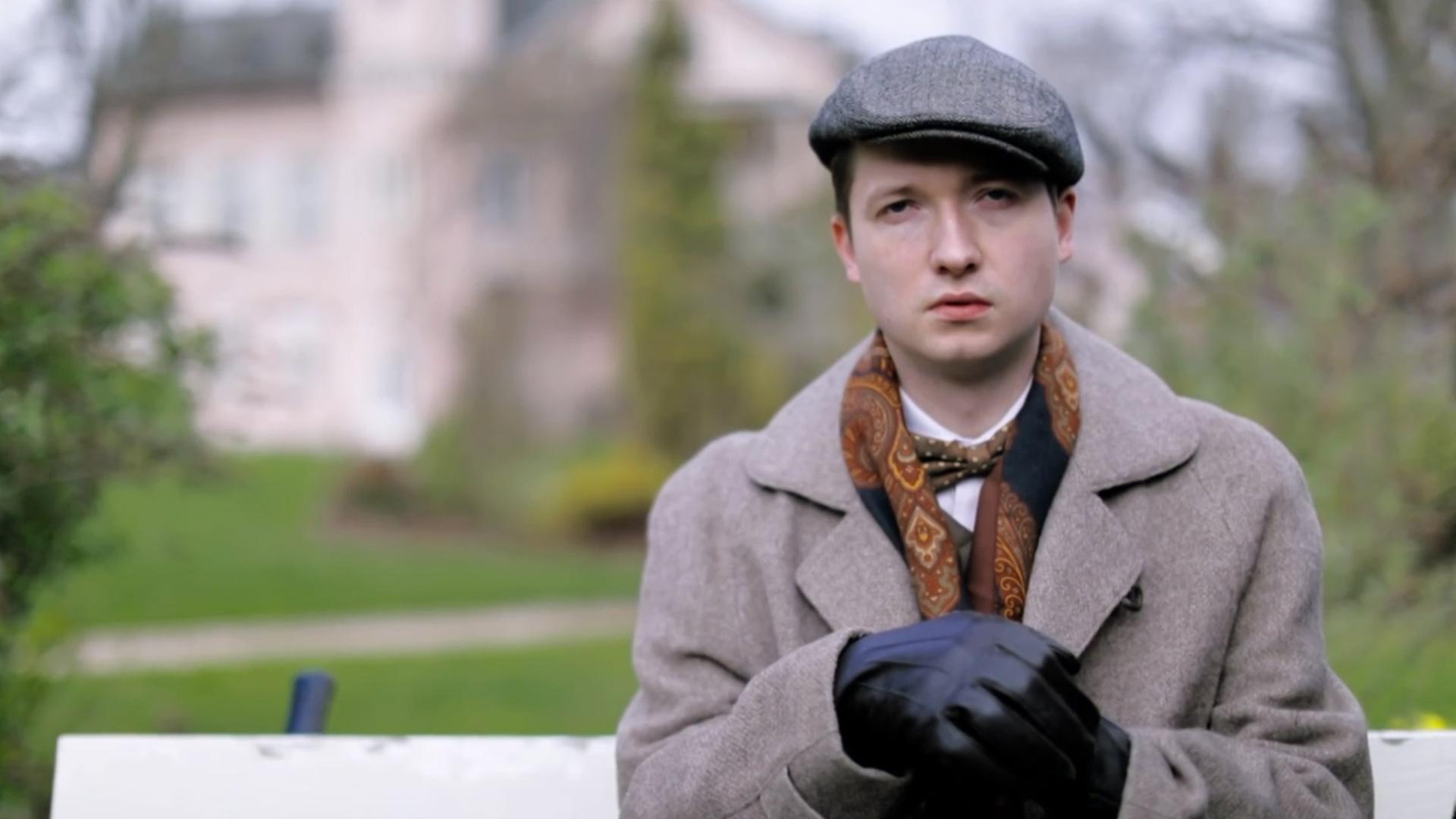 Szene aus "Empusion" von Olga Tokarczuk: Ein junger Mann, gekleidet mit grauer Baskenmütze, Mantel, Schal und dunklen Lederhandschuhen, sitzt auf einer weißen Parkbank. Seine Hände liegen übereinander gefaltet auf einem Stock.