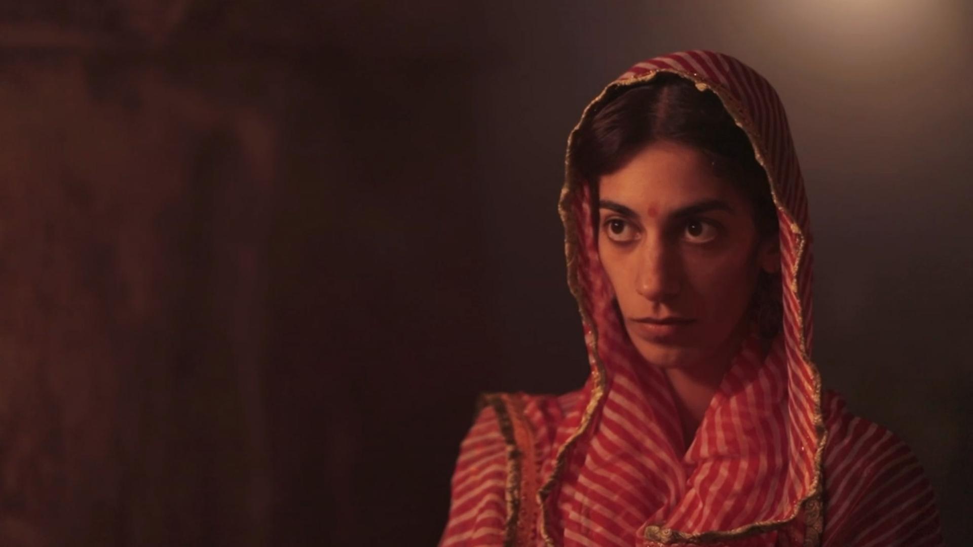 Szene aus "Victory City" von Salman Rushdie: Junge Frau mit einem rot-weiß gemusterten Kopftuch steht vor einer dunklen Wand.