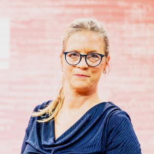 Eine Person mit Brille steht vor einem rosa Hintergrund mit dem weißen Schriftzug „BUCHZEIT“. Sie trägt ein dunkelblaues Oberteil. Das Porträt entstand im Rahmen der Frankfurter Buchmesse 2025 während der Aufzeichnung der Literatursendung „Buchzeit“ auf der Bühne von ARD, ZDF und 3sat.