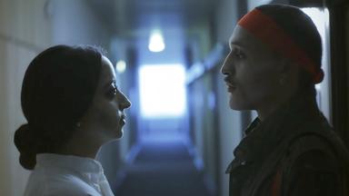 Szene aus "Die sieben Monde des Maali Almeida" von Shehan Karunatilaka. Seitliche Ansicht von einer Frau und einem Mann, die in einem langen düsteren Gang stehen und sich ansehen.