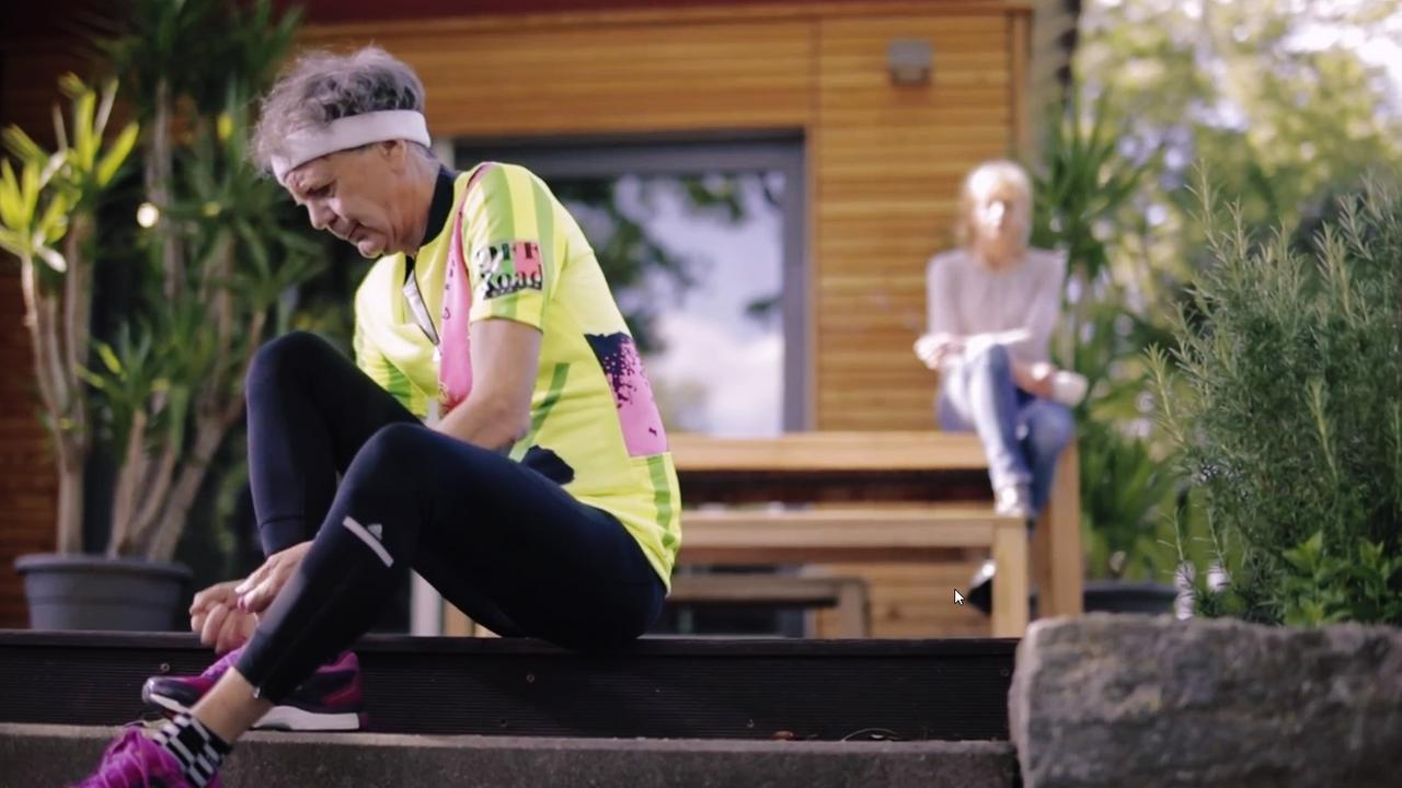 Szene aus "Die Letzten werden die Ersten sein" von Lionel Shriver. Ein älterer Mann mit grauen Haaren in Sportbekleidung sitzt auf einer Treppenstufe einer Holzterrasse und bindet sich seinen Sportschuh. Im Hintergrund sitzt eine ältere, nachdenkliche Frau auf einem Holzgartentisch.