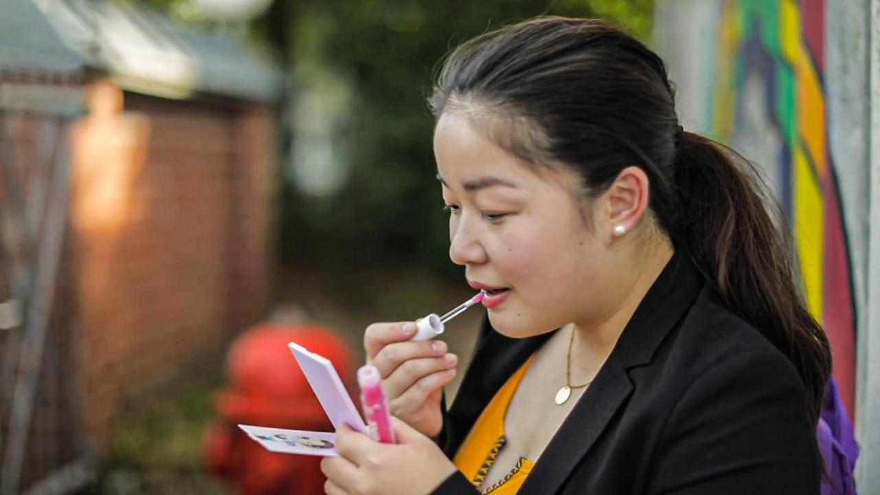 Szene aus "Die kleinen Lügen der Ivy Lyn" von Susie Yang. Eine junge Frau schminkt sich die Lippen mit einem pinken Lippenstift. In der linken Hand hält sie einen Spiegel.
