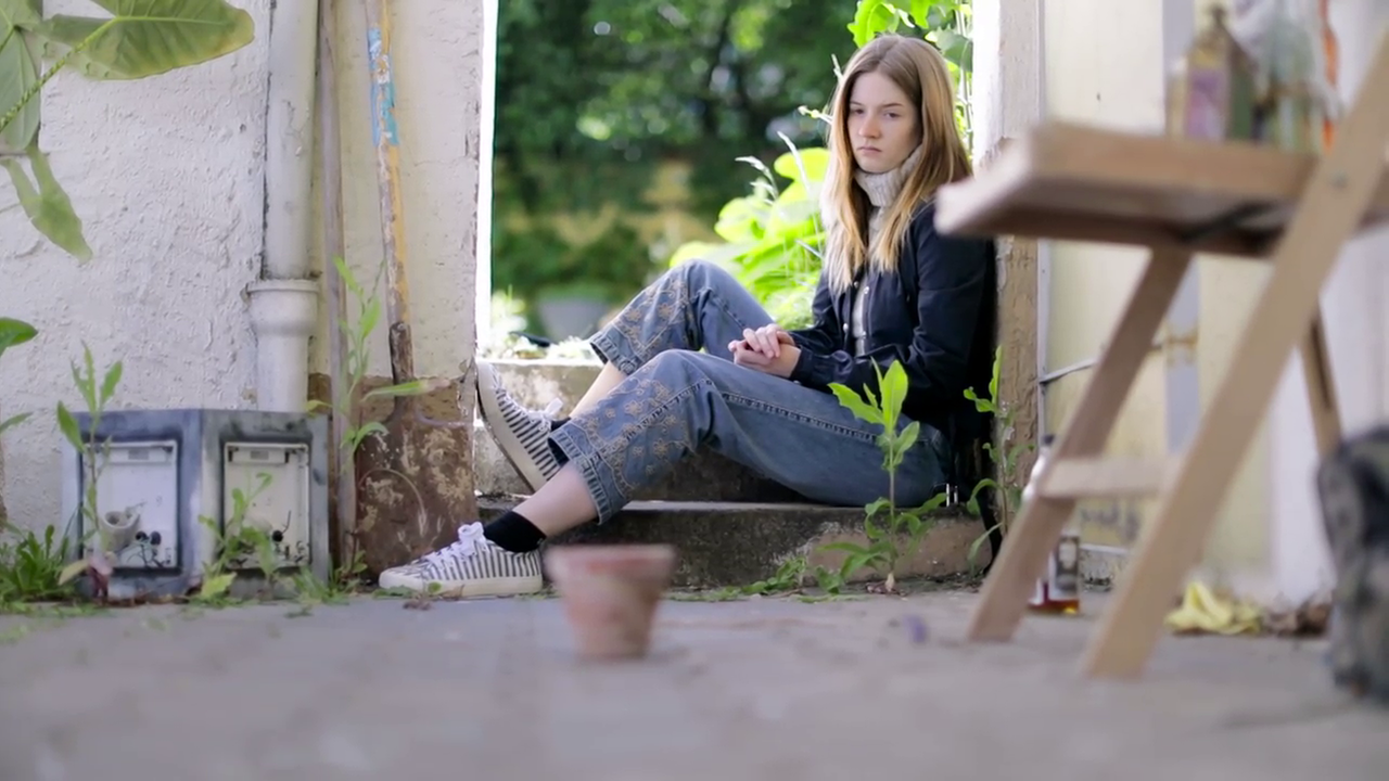 Szene aus "Der Garten aus Glas" von Tatiana Țîbuleac: Eine junge Frau sitzt auf einer Stufe in einem Türrahmen im Freien. In der Mitte vor ihr steht ein alter Tonblumentopf, den sie deprimiert ansieht. Rechts von ihr steht eine halb leere Flasche auf dem betonierten Boden des Hofes. 