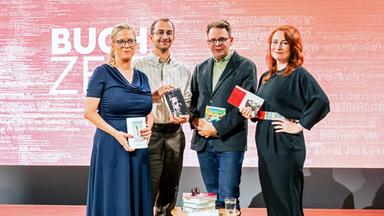 Vier Personen stehen auf einer Bühne mit pink-rotem Hintergrund, auf dem groß „BUCHZEIT“ zu lesen ist. Jede Person hält ein Buch in der Hand. Vor ihnen steh ein kleiner Tische mit Bücherstapeln. Auf der linken Seite steht ein zweiter kleiner Tisch mit einem Bücherstapel und einem Glas Wasser. Links neben einem Tisch steht ein Lautsprecher und der Bühnenboden ist aus Holz.  