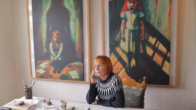 Szene aus "Die Wahrheiten meiner Mutter" von Vigdis Hjorth. Ein Frau mit roten Haaten sitzt an einem Tisch und telefoniert mit ihrem Handy. Im Hintergrund hängen zwei gerahmte Gemälde. Die Bilder zeigen jeweils ein junges, rothaariges Mädchen in einem Kleid. Die Farben der Bilder sind dunkel und die Bilder wirken düster. Auf dem einen Bild ist hinter dem Mädchen ein dunkler, großer Schatten. Das Mädchen auf dem anderen Gemälde schreit.  