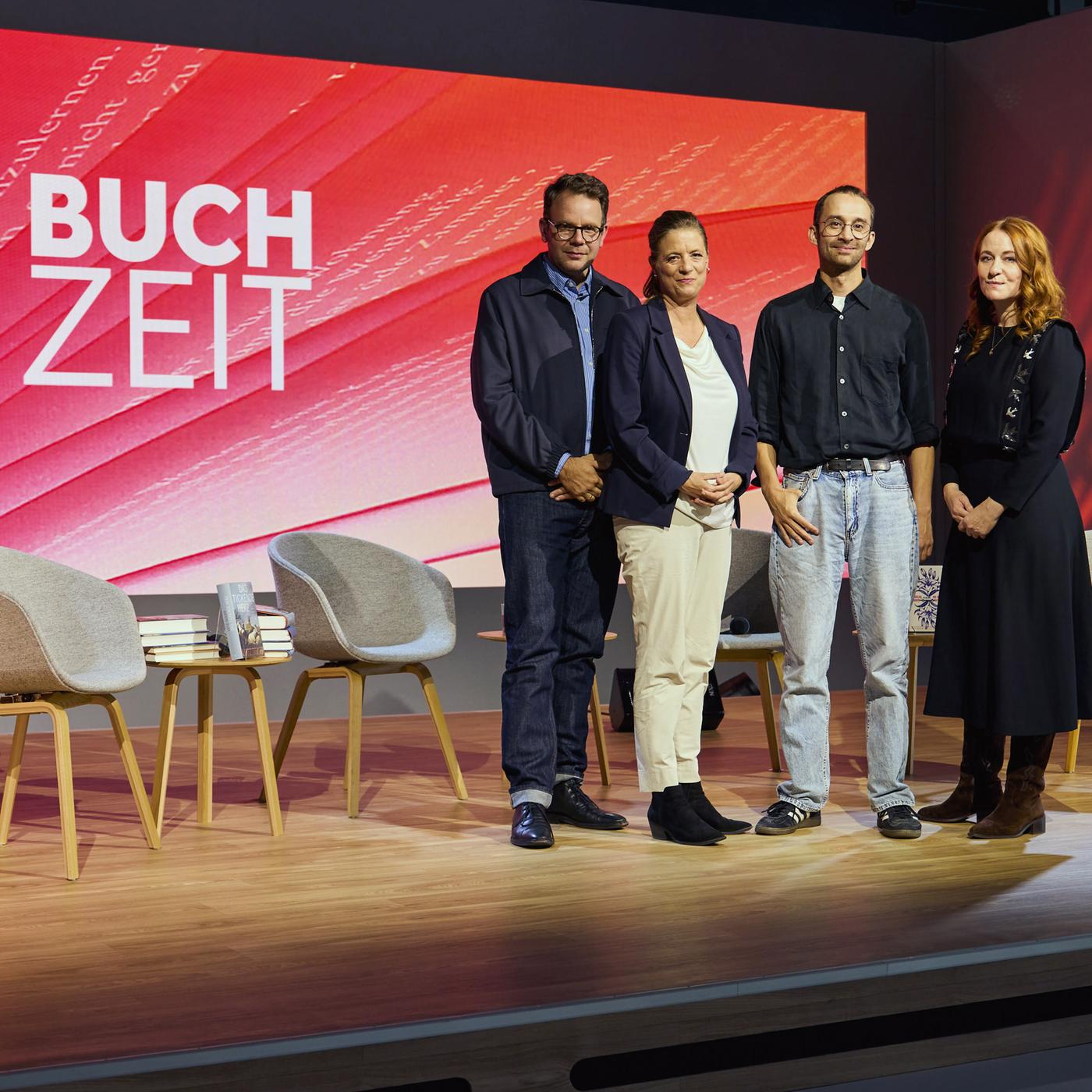 Gruppenfoto auf der Bühne der Literatursendung „Buchzeit“: Vier Personen posieren vor einer rot gestalteten Hintergrundwand mit Sendungslogo. Im Vordergrund stehen Stühle und Bücher.