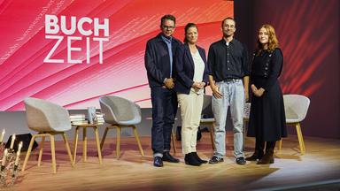 Gruppenfoto auf der Bühne der Literatursendung „Buchzeit“: Vier Personen posieren vor einer rot gestalteten Hintergrundwand mit Sendungslogo. Im Vordergrund stehen Stühle und Bücher.