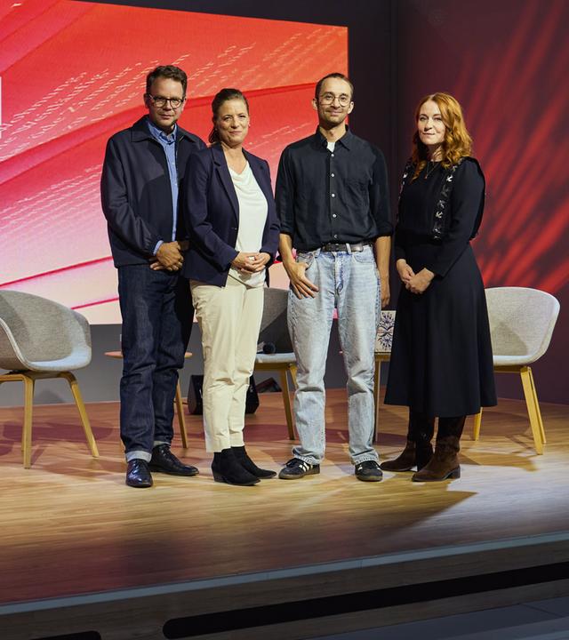 Gruppenfoto auf der Bühne der Literatursendung „Buchzeit“: Vier Personen posieren vor einer rot gestalteten Hintergrundwand mit Sendungslogo. Im Vordergrund stehen Stühle und Bücher.