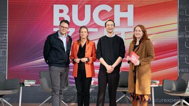 Gruppenfoto auf einer Studiobühne der Sendung „Buchzeit“. Vier Personen stehen auf einer beleuchteten Bühne mit dem Schriftzug „BUCHZEIT“ im Hintergrund; umgeben sind sie von mehreren Sesseln und Büchern als Teil des Bühnenbilds. 