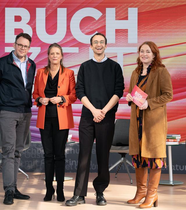 Gruppenfoto auf einer Studiobühne der Sendung „Buchzeit“. Vier Personen stehen auf einer beleuchteten Bühne mit dem Schriftzug „BUCHZEIT“ im Hintergrund; umgeben sind sie von mehreren Sesseln und Büchern als Teil des Bühnenbilds. 