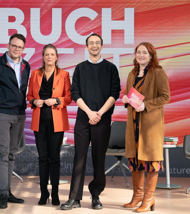 Gruppenfoto auf einer Studiobühne der Sendung „Buchzeit“. Vier Personen stehen auf einer beleuchteten Bühne mit dem Schriftzug „BUCHZEIT“ im Hintergrund; umgeben sind sie von mehreren Sesseln und Büchern als Teil des Bühnenbilds. 