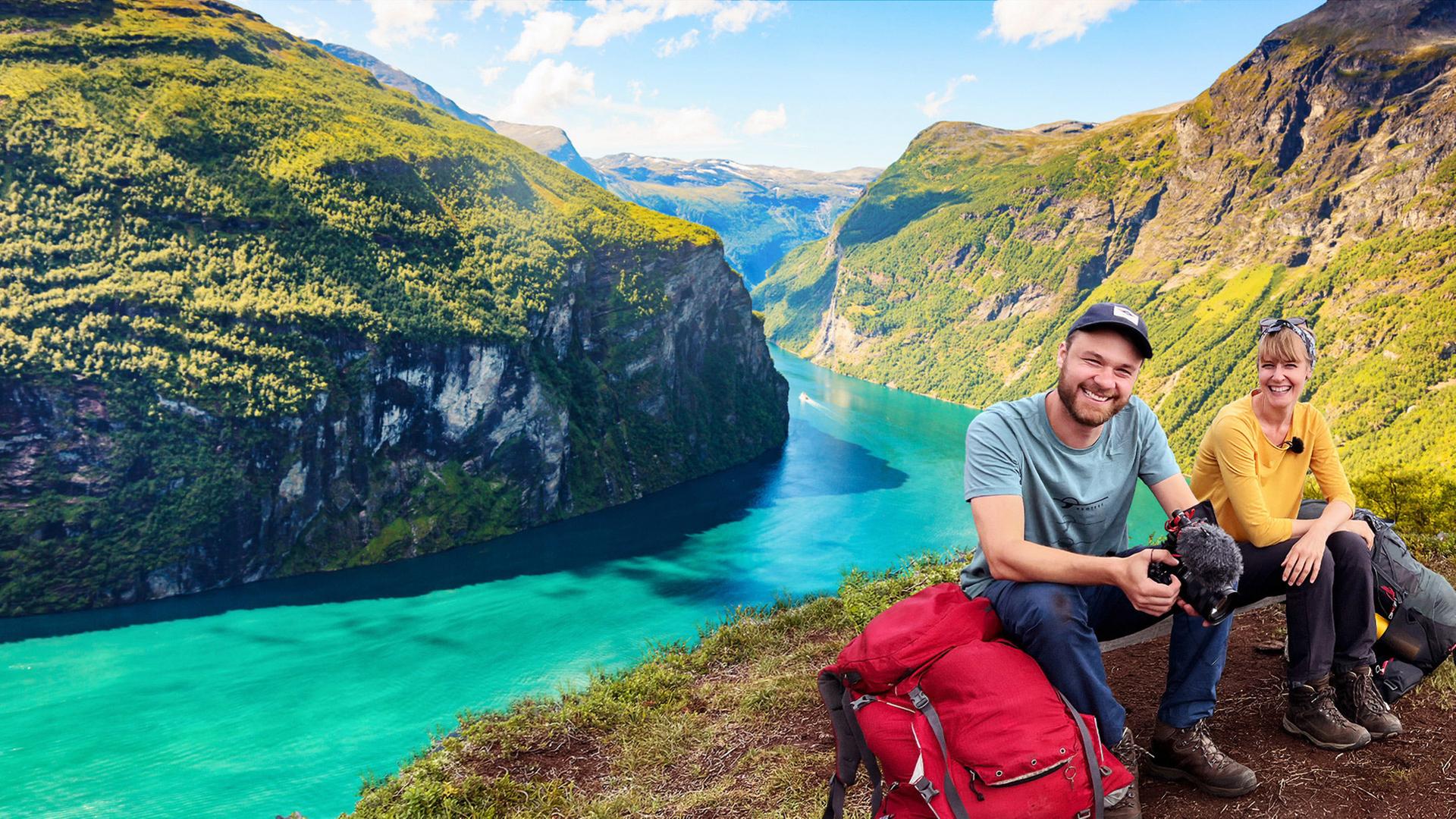 Ein Mann mit Kamera in der Hand und eine Frau sitzen hoch über einem norwegischen Fjord, dessen Wasser türkis ist.